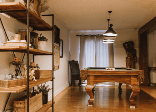 A cozy game room featuring a pool table, wooden shelves with decor, and soft lighting from pendant lamps.