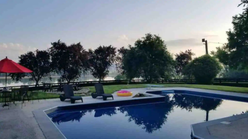 A serene poolside scene at dawn, featuring lounge chairs, a colorful float, and trees reflecting in the water.