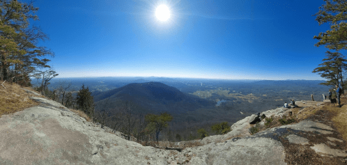 Panoramic view from a rocky overlook, showcasing rolling hills and a bright sun in a clear blue sky.