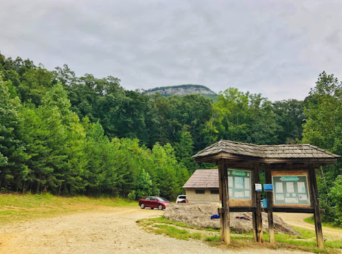 A gravel path leads to a wooden information kiosk surrounded by lush green trees and a mountain in the background.