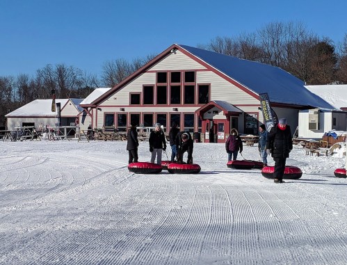 A snowy scene with people preparing to tube outside a ski lodge on a clear day.