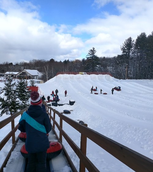 A snowy hill with people tubing, surrounded by trees and a lodge in the background under a partly cloudy sky.