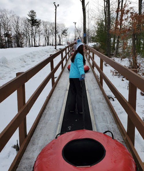 A person in a blue jacket pulls a red snow tube up a snowy ramp surrounded by trees.