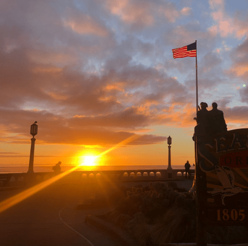 A sunset over the ocean with a flag waving and a statue in the foreground, casting a warm glow on the scene.
