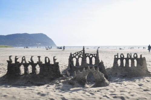 Sandcastles on a beach with "SEASIDE" and "HONO" carved into them, against a backdrop of people and mountains.