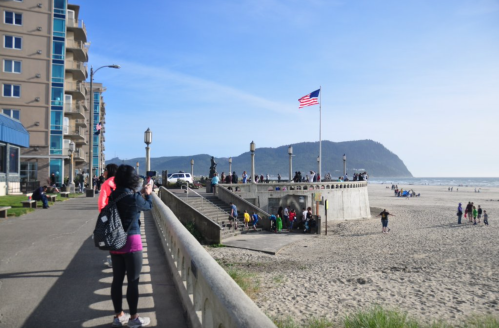 A beachside scene with people walking, taking photos, and a flag flying, set against a backdrop of hills and blue sky.