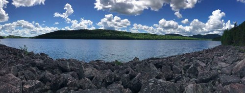 A panoramic view of a serene lake surrounded by lush green hills and a blue sky filled with fluffy white clouds.