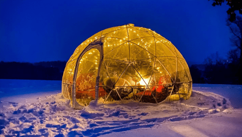 A glowing geodesic dome in a snowy landscape, illuminated with warm lights against a dark blue evening sky.