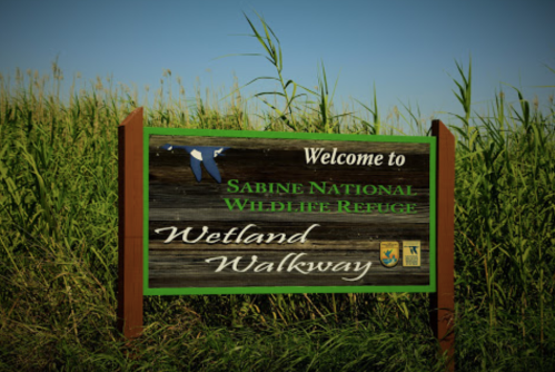 Sign welcoming visitors to Sabine National Wildlife Refuge, featuring "Wetland Walkway" and surrounded by tall grass.