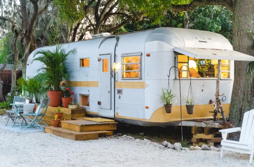 A vintage silver trailer surrounded by greenery, with potted plants and outdoor seating on a gravel path.