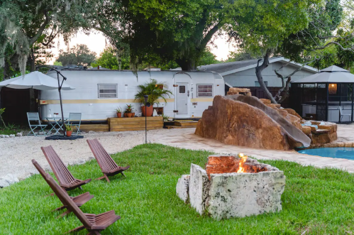 A cozy outdoor space featuring a vintage trailer, fire pit, and lounge chairs surrounded by greenery and a pool.