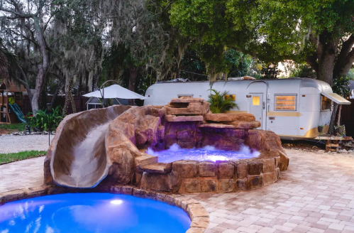 A backyard scene featuring a stone water slide, a pool with blue water, and an Airstream trailer in the background.