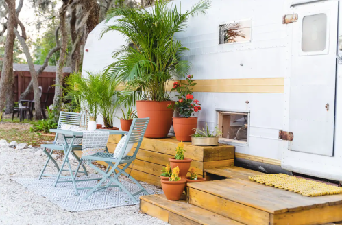 A cozy outdoor space with a vintage trailer, potted plants, a small table, and chairs on wooden steps.