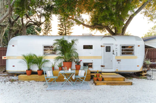 A vintage trailer surrounded by greenery, with a cozy outdoor seating area and potted plants on a gravel surface.