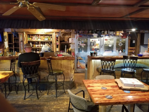 A cozy bar interior with wooden furniture, a bartender in the background, and a customer sitting at the counter.