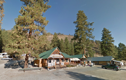 A rustic lodge surrounded by tall pine trees, with a gravel parking area and mountains in the background.