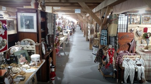 A spacious indoor market with wooden beams, showcasing various booths filled with antiques and crafts along a long aisle.