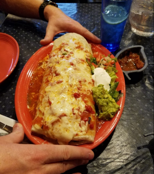 A large burrito topped with cheese and sauce, served with guacamole, sour cream, and a side of salsa on a red plate.