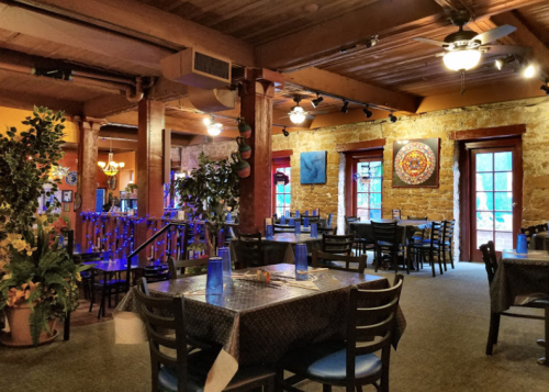 Cozy restaurant interior with stone walls, wooden beams, and tables set for dining, featuring colorful artwork and plants.