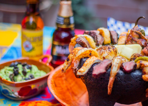A vibrant table setting featuring a colorful dish of grilled skewers, guacamole, and two bottles of beer.