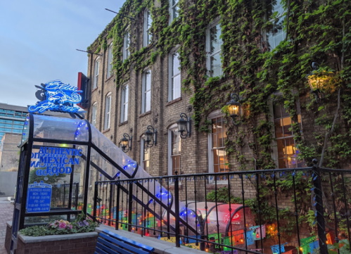 A charming brick building covered in ivy, featuring a neon sign and colorful decorations outside a Mexican restaurant.