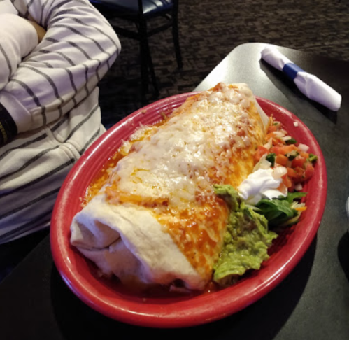 A large burrito topped with melted cheese, served with guacamole, sour cream, and fresh tomatoes on a red plate.