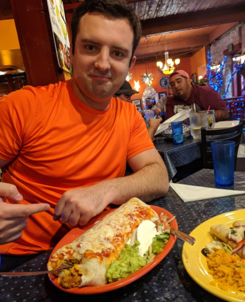 A man in an orange shirt points at a large burrito on a plate, with guacamole and sour cream, in a colorful restaurant.