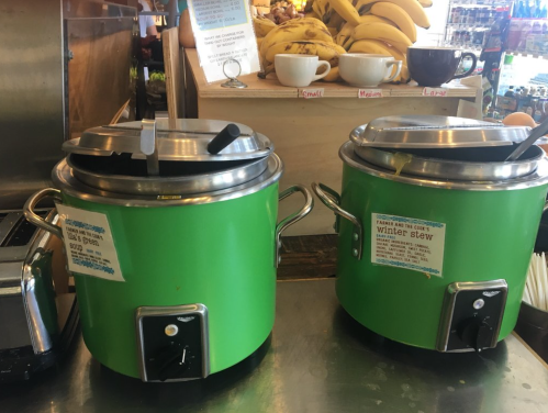 Two green soup kettles on a counter, labeled "Winter stew," with utensils resting on top. Bananas and cups in the background.