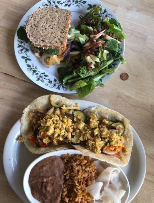 A plate with two tacos filled with vegetables and crumbled topping, alongside rice, beans, and a side salad.