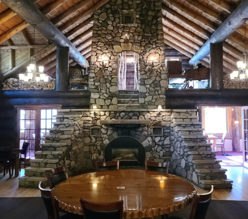 Cozy lodge interior featuring a stone fireplace, wooden beams, and a round dining table. Warm, rustic atmosphere.