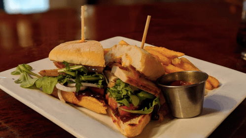 A sandwich with grilled chicken, greens, and sauce, served with fries and a small cup of ketchup on a white plate.