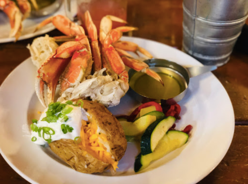 A plate featuring crab legs, a baked potato with toppings, and a side of mixed vegetables, with a dipping sauce.