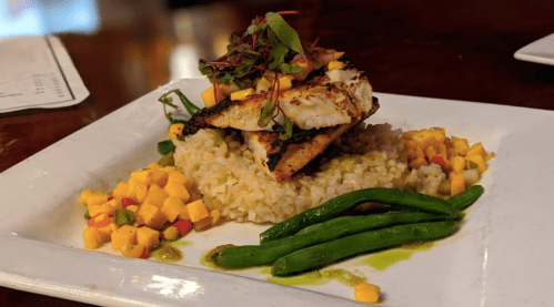 A plate of grilled fish atop rice, garnished with mango, green beans, and microgreens.