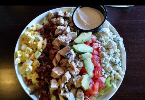A colorful salad bowl with grilled chicken, bacon, eggs, tomatoes, avocado, and a side of dressing.