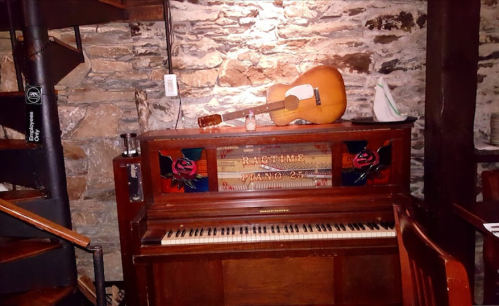 A vintage piano labeled "Ragtime Piano 25" sits against a stone wall, with a guitar resting on top.
