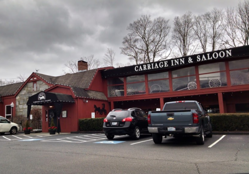 Exterior of Carriage Inn & Saloon, featuring a red building, large windows, and parked cars in front. Overcast sky.