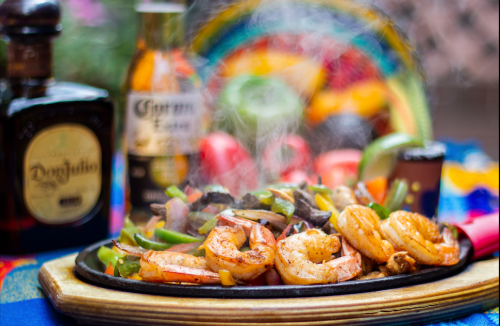Sizzling shrimp fajitas on a wooden platter, surrounded by colorful peppers, with drinks in the background.