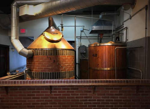 Two large brewing kettles, one copper and one wooden, in a brewery setting with exposed pipes and brick walls.