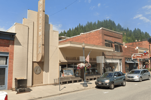 Historic storefronts line a street, featuring a museum, flower baskets, and an American flag against a blue sky.