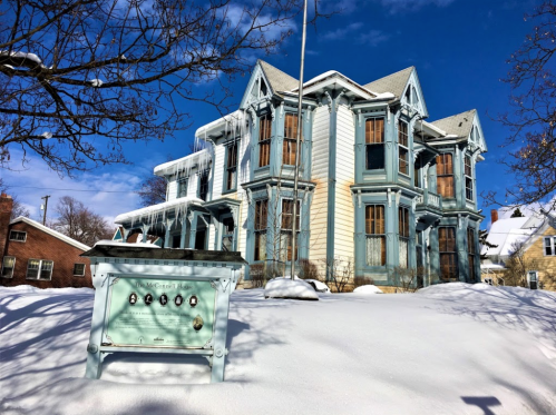A large, historic blue house with snow-covered grounds and a sign in front, under a clear blue sky.