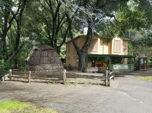 A rustic building with a thatched hut nearby, surrounded by trees and a wooden fence.