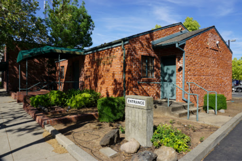 A brick building with a green awning, entrance sign, and landscaped area, surrounded by trees and parking.