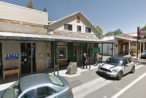 A street view of a small town with shops, people walking, and parked cars under a clear blue sky.