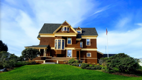 A large, brown Victorian-style house with a manicured lawn and blue sky in the background. An American flag is visible.