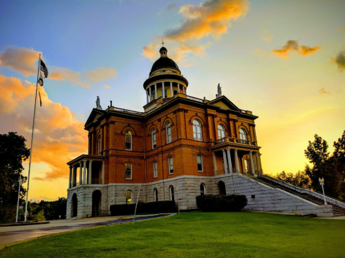 Historic brick building with a dome, surrounded by greenery, under a colorful sunset sky. American flag in the foreground.