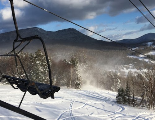 A ski lift chair hangs over a snowy landscape with mountains and trees, under a partly cloudy sky.