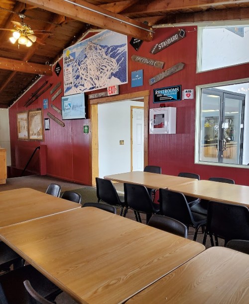 Interior of a rustic room with wooden tables, chairs, and various signs on the walls, including maps and restroom information.