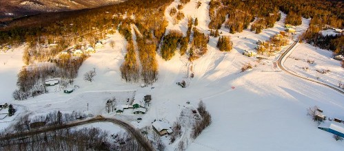Aerial view of a snowy landscape with houses, trees, and winding roads under sunlight.