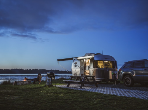 A silver Airstream trailer by a lake at dusk, with a picnic table, fire pit, and a car parked nearby.