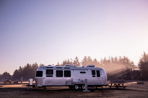 A silver Airstream trailer parked in a misty campsite with trees in the background and soft morning light.
